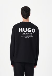 Black sweatshirt featuring a large white "HUGO" logo and text on the back, with a relaxed fit and long sleeves. Soft cotton fabric.
