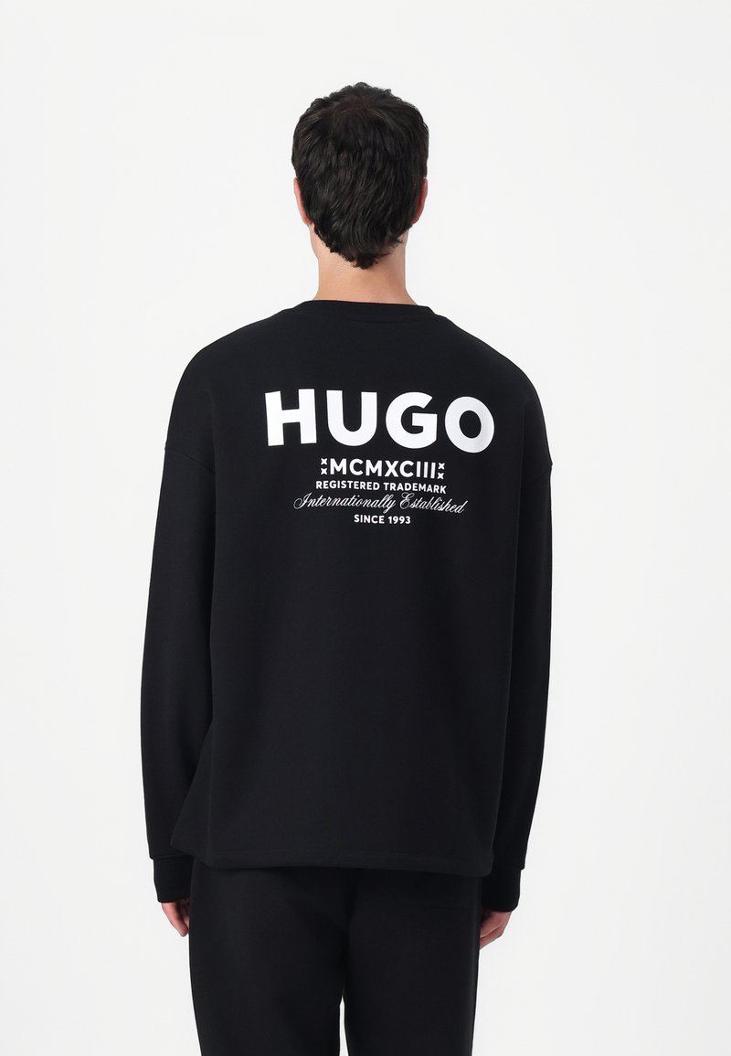 Black sweatshirt featuring a large white "HUGO" logo and text on the back, with a relaxed fit and long sleeves. Soft cotton fabric.
