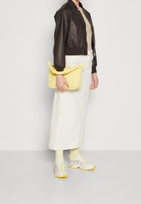 Hnědá bomber bunda přes béžový top, bílou sukni a světle žluté doplňky, včetně volánkového psaníčka a tenisek se žlutými akcenty.