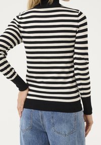 Personne portant un t-shirt à manches longues à rayures horizontales noires et blanches et un jean en denim bleu, vue de dos avec une main sur la hanche.