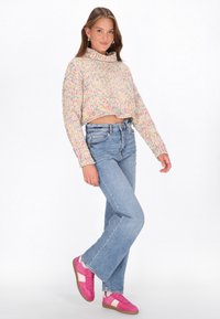Gehaakte, pastelgekleurde trui met een hoge kraag; gecombineerd met lichtblauwe jeans en roze sneakers met witte accenten.