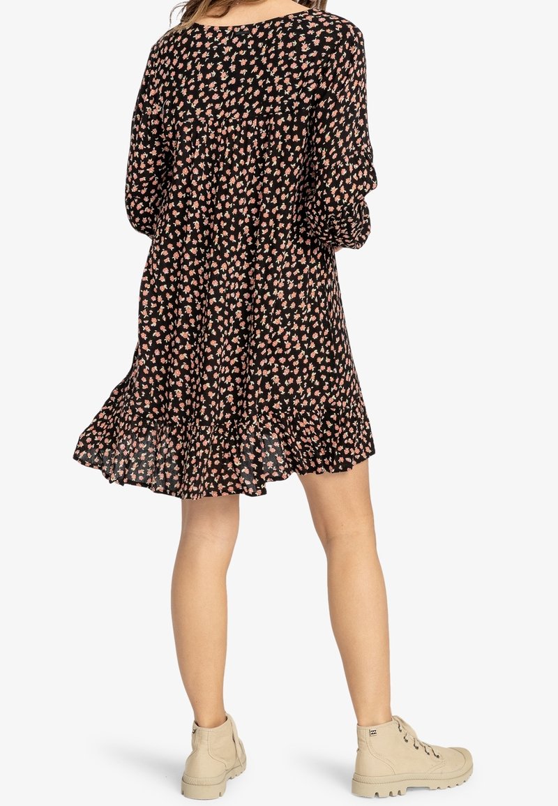 Femme portant une robe noire à imprimé floral avec manches longues et ourlet à volants, associée à des bottines beige à lacets, debout, dos à l'avant.
