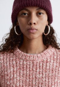 Rosa und weiße strukturierte Strickjacke mit rundem Ausschnitt, kombiniert mit einer dunkelroten gerippten Strickmütze und großen silbernen Creolen.