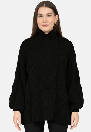 Junge Frau mit langem braunem Haar, die einen schwarzen, strukturierten, kabelgestrickten Rollkragenpullover und goldene Creolen trägt, blickt nach vorne.