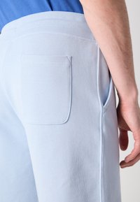 Personne portant un pantalon de survêtement bleu clair avec poche arrière et couture latérale, debout avec la main droite détendue le long du corps.