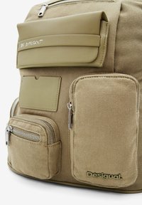 Béžový batoh s texturovanou tkaninou, několika kapsami na zip a přední klopou s nápisem „BUĎ JINÝ“. Logo je vyraženo na spodní části.