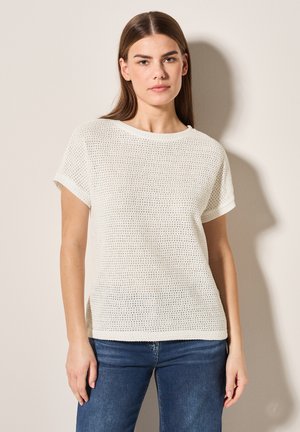 Femme aux cheveux bruns raides portant un haut texturé blanc à manches courtes et un jean bleu, debout contre un mur clair.