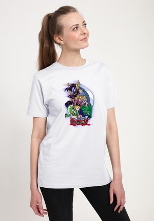 Kvinde med hestehale iført hvid T-shirt med farverige Yu-Gi-Oh-figurer og logo, der kigger op og smiler let.