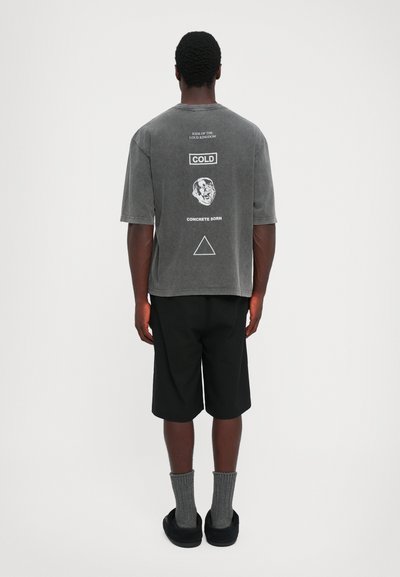 Man met zijn rug naar de camera, gekleed in een vervaagde grijze T-shirt met witte graphics en tekst, zwarte shorts, grijze sokken en zwarte instap schoenen.