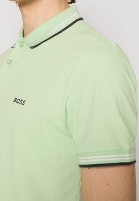 Homme portant un polo BOSS vert clair avec un col rayé et des bords de manches rayés, boutonné au niveau du cou.