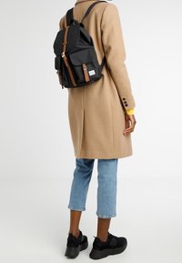 Zwarte rugzak met bruine leren accenten, twee voorzakken, trekkoordsluiting en verstelbare riemen. Draagt deze op een persoon met een beige jas.