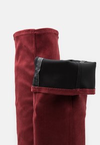 Burgundy knähöga stövlar med en mjuk mockayta, som har en svart tygkrage och kontrastrikningar som framhäver de unika designelementen.