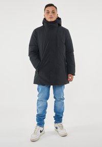 Parka nera imbottita con colletto alto, maniche lunghe, chiusura frontale con zip e tasche laterali. Indossata sopra jeans blu e sneaker bianche.