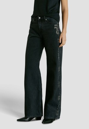 Mujer vistiendo vaqueros negros de pierna ancha con adornos de tachuelas plateadas a lo largo de las costuras exteriores y botas negras de punta al tobillo.