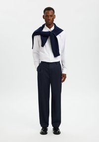 Λευκό πουκάμισο με κουμπιά, μπλε σκούρο πουλόβερ ριγμένο στους ώμους, σκούρο παντελόνι, μαύρα παπούτσια. Το μοντέλο στέκεται μπροστά σε απλό ανοιχτό φόντο.