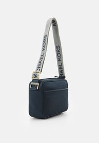 Granatowa torebka na ramię o gładkiej fakturze, z metalowym zapięciem w kolorze złotym i paskiem z szarym logo "MICHAEL KORS". Prostokątny kształt.