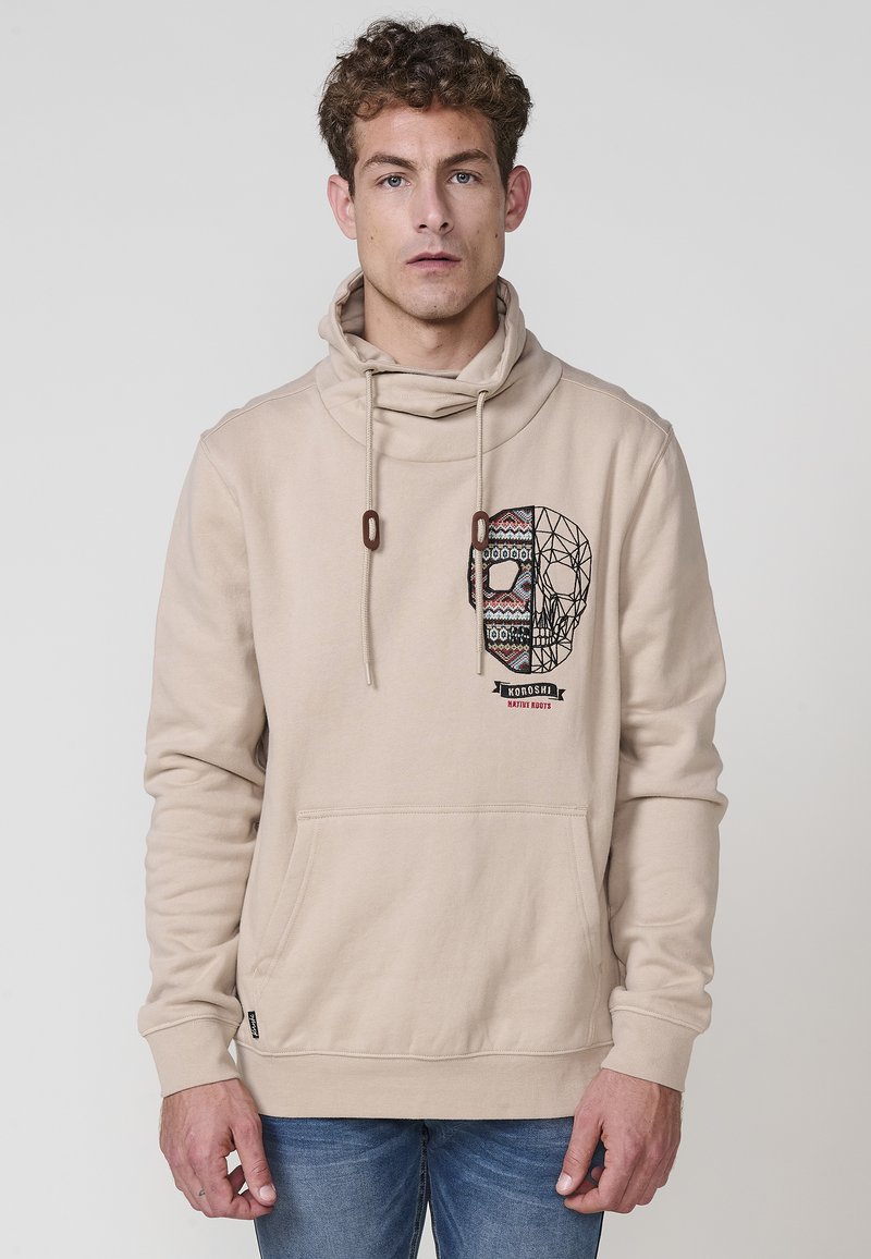 Beige hoodie med krage och dragsko. Har en färgglad dödskallegrafik och framficka. Tillverkad av mjukt, texturerat tyg.