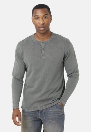 Hombre con una camiseta henley de manga larga gris y vaqueros azules, de pie con una mano en el bolsillo contra un fondo liso.
