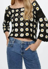 Zwart gehaakte trui met een patroon van witte en gele madeliefjes; heeft een brede halslijn en driekwart mouwen, gecombineerd met een blauwe spijkerbroek.