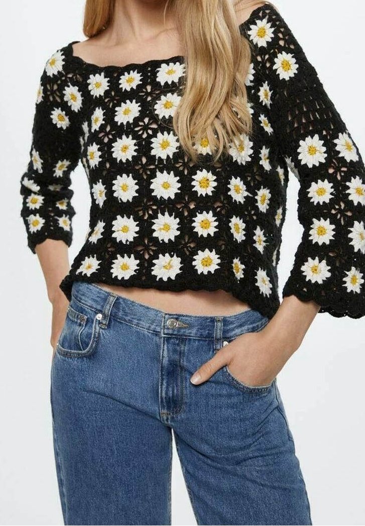 Zwart gehaakte trui met een patroon van witte en gele madeliefjes; heeft een brede halslijn en driekwart mouwen, gecombineerd met een blauwe spijkerbroek.