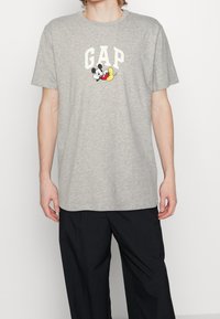 Grå bomulds t-shirt med et hvidt "GAP" logo og farverig Mickey Mouse grafik. Korte ærmer, rund halsudskæring, standard pasform.