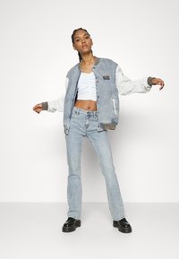 Denim bomberjack met witte mouwen, ribgebreide manchetten en "GUESS" borduursel. Draag het over een witte crop top met rimpeldetails en lichtblauwe spijkerbroeken.