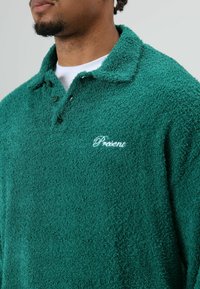 Groene, pluizige trui met kraag en een getextureerde fleece stof. Voorzien van een twee-knoppen plooi en geborduurd woord "Present" in wit op de borst.