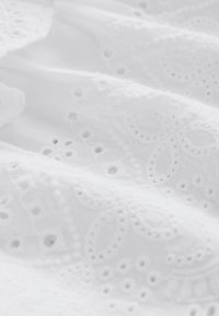 Valge kangas, millel on keerukad tikitud mustrid, silmaaugudetailid ja pehme tekstuur. Disain sisaldab lille- ja ringikujulisi motiive.