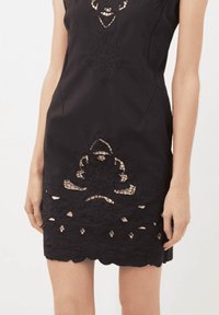 Vestido negro sin mangas con recortes de encaje floral en el pecho, la parte media y el dobladillo, usado por una persona de pie con los brazos relajados.