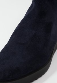 Tmavě modrá semišová kotníková bota s hladkým povrchem, elegantním designem a silnou černou podrážkou, zdůrazňující minimalistické prvky.