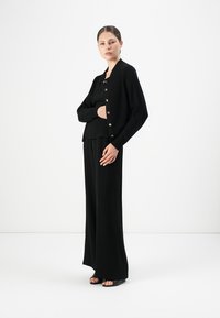 Woman standing wearing black buttoned cardigan, black top, wide-leg pants, and black open-toe sandals against a plain white background.