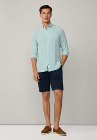 Camisa de lino azul claro con colla de botones, mangas enrolladas; combinada con pantalones cortos navy y zapatos de deslizamiento marrones, sobre un fondo neutro.