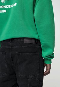 Persona che indossa una felpa verde con testo bianco e jeans neri di denim con grandi tasche posteriori, mano rilassata di lato.