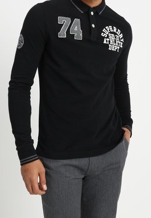 Homme portant un polo noir à manches longues avec le numéro 74 et le texte "Superdry Athletic Dept", associé à un pantalon gris.