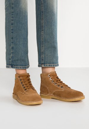 Bottes chelsea en suède marron avec des lacets robustes, œillets en argent, une semelle claire contrastante et un design à bout rond. Portées avec un jean en denim bleu.