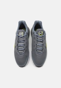 Zapatillas deportivas de malla gris con un logotipo negro redondo, acentos verdes y suela texturizada. Presentan cordones con ojales reforzados para un ajuste seguro.