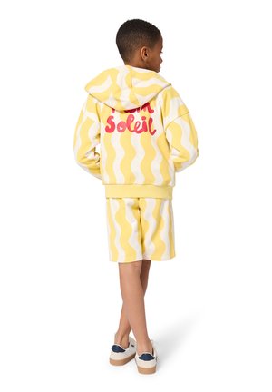 Enfant portant un sweat à capuche jaune et blanc à motif ondulé et un short, texte rouge au dos du sweat, debout de dos, baskets blanches.