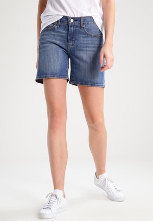 Persoon draagt mid-lange blauwe denim shorts, witte sneakers en een ingestopt wit t-shirt, staand tegen een effen achtergrond.