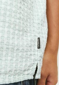 Chemise texturée bleu clair avec un motif gaufré, fente sur le côté, et une étiquette noire portant la mention "ORIGINALIST" attachée près de l'ourlet.
