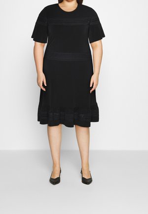 Kobieta plus size w czarnej sukience do kolan z krótkimi rękawami, ozdobnym poziomym przeszyciem i czarnych butach na szpiczastym obcasie, stojąca na tle białej ściany.