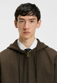 Bruine herringbone zip-up hoodie met capuchon, trekkoord, gouden rits en een witte coll shirt eronder, gecombineerd met een zwarte stropdas.