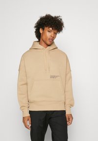 Felpa beige con tasca a kangaroo e cappuccio regolabile. Presenta un logo stampato in nero sul petto. Tessuto morbido, vestibilità comoda.