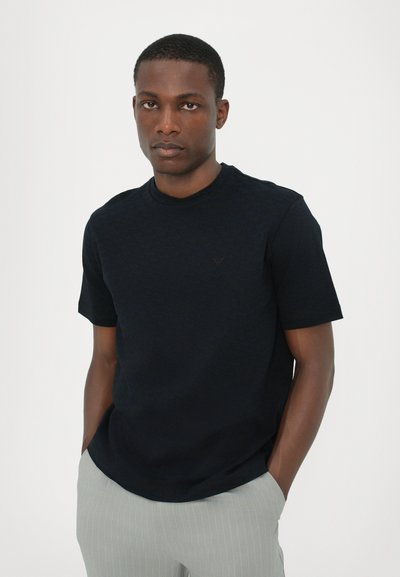 T-shirt noir à manches courtes avec un motif texturé, encolure ronde et un détail de logo subtil. Associé à un pantalon gris clair à rayures.