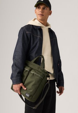 Sac vert olive avec plusieurs poches, fabriqué à partir d'un tissu lisse. Il dispose de poignées doubles et d'une sangle détachable. Porté par une personne en veste en jean.