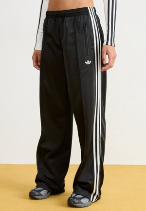Persona vistiendo pantalones anchos negros de Adidas con rayas blancas, bolsillo lateral con cremallera y zapatillas grises sobre un suelo amarillo.