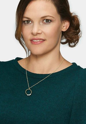 Goldene Halskette mit einem runden Anhänger, der drei kleine weiße Perlen enthält. Das Model trägt einen dunkelgrünen Pullover mit einer strukturierten Oberfläche.