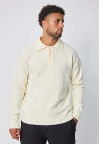 Pull en tricot de couleur crème avec un col polo, quatre boutons, des poignets et un ourlet côtes. Texture douce et coupe décontractée.