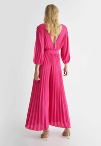 Abito maxi plissettato di un vivace rosa con profonde aperture a V sulla schiena, maniche lunghe a palloncino e vita cinte. Presenta un tessuto testurizzato e una silhouette fluida.