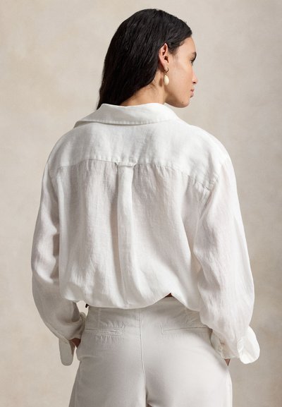 Vrouw met lang donker haar draagt een losse witte linnen blouse en witte broek, staand tegen een neutrale achtergrond, gezien van achteren en vanaf de zijkant.