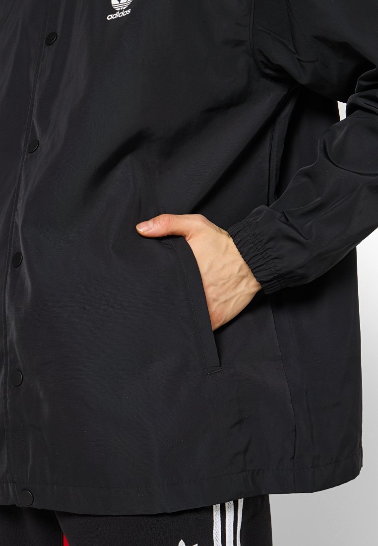 Hand in die Seitentasche einer schwarzen Adidas-Jacke mit elastischem Bündchen und Logo auf der Brust, das einen Teil der Adidas-Hose zeigt.